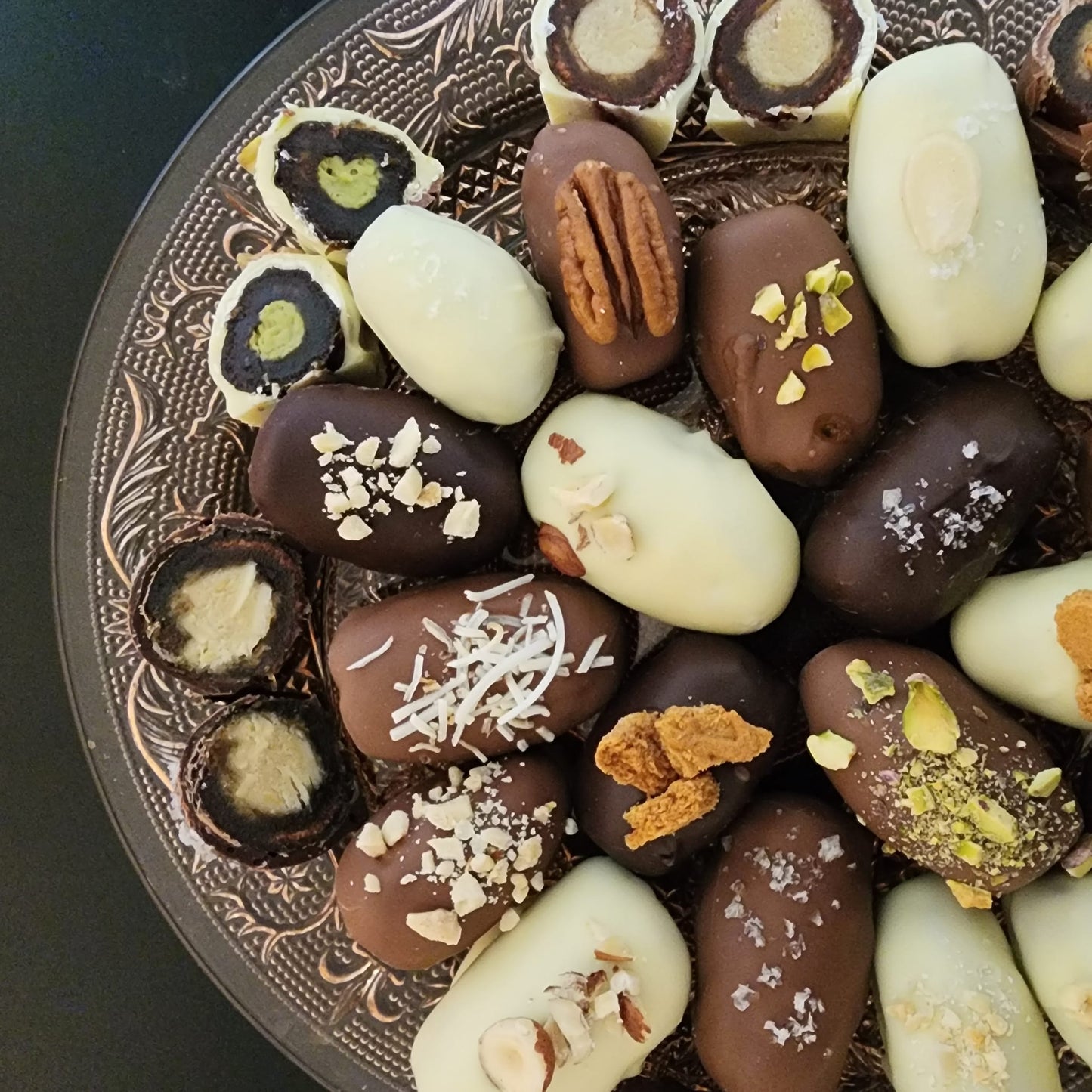 Close view of assorted chocolate-covered dates on a patterned glass plate, topped with pecans, chopped hazelnuts, coconut and pistachio, cut pieces revealing marzipan and pistachio fillings