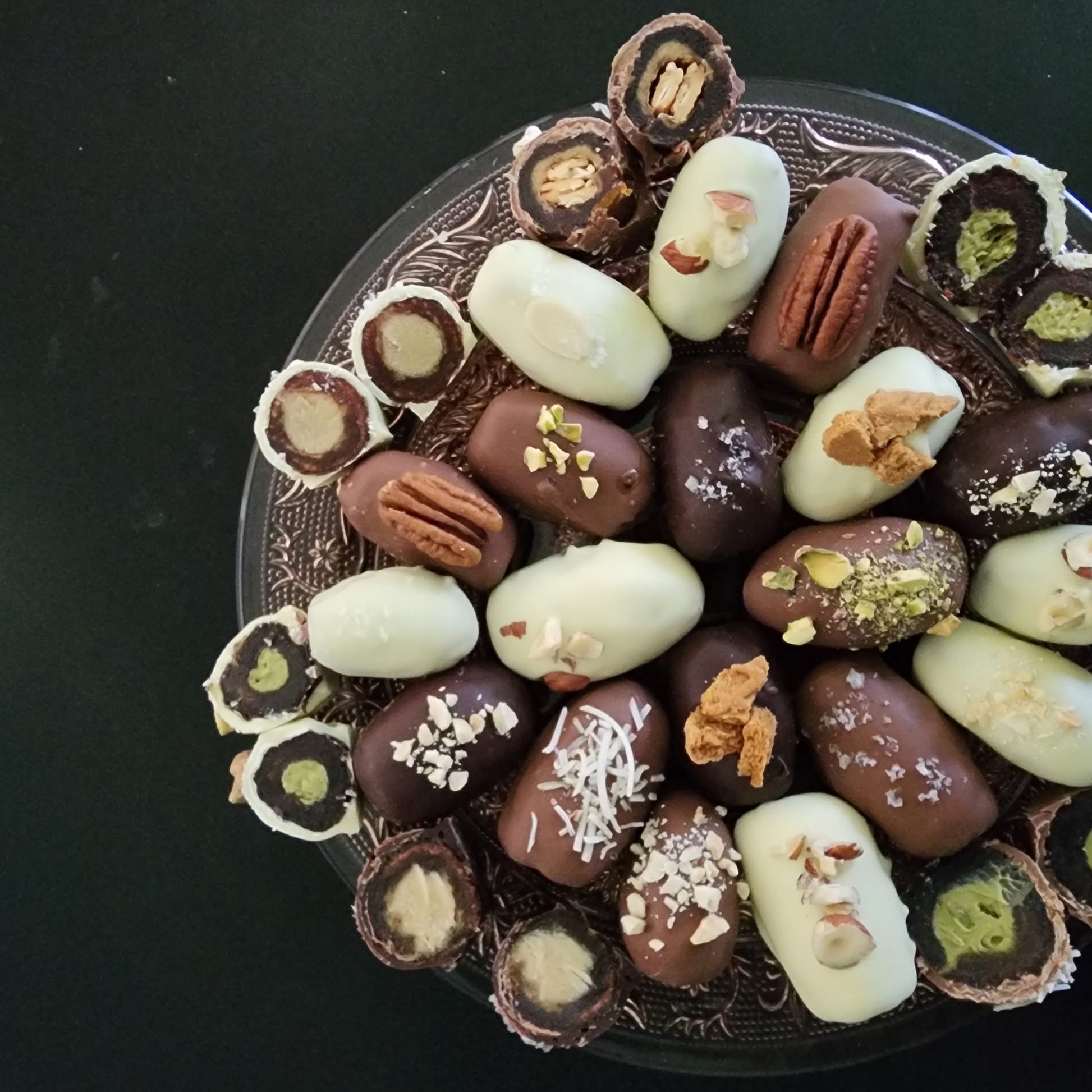 Platter of assorted chocolate-enrobed Medjool dates showing dark, milk and white chocolate varieties with pecan, pistachio, coconut and sea-salt toppings, several halved to show pistachio and almond-butter centres