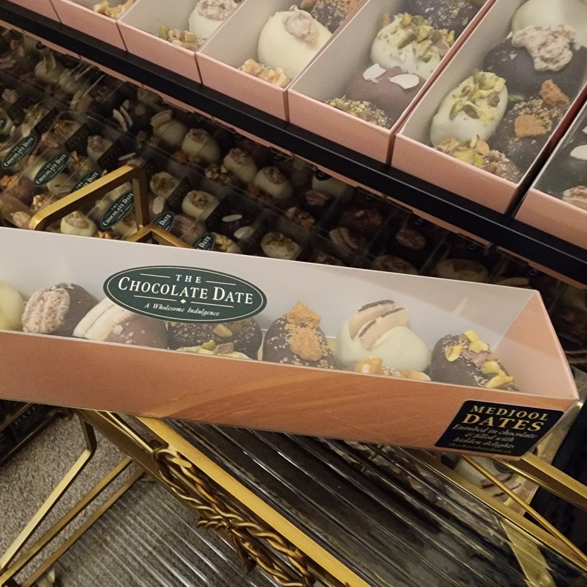 Long 900 gram gift box of assorted chocolate-covered Medjool dates with a clear lid and The Chocolate Date oval label, displayed at a market stall